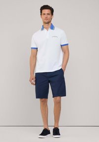 Biała koszulka polo z niebieskim kołnierzykiem i akcentami na rękawach, w połączeniu z granatowymi szortami i czarnymi sneakersami. Prosty, strukturalny design z detalem logo.