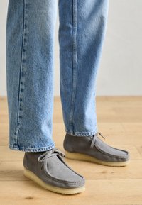 Scarpe in suede grigio con punta rotonda, suola piatta in gomma e design con lacci, indossate con jeans in denim azzurro chiaro su un pavimento di legno.