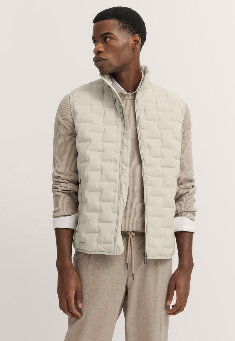 Helle beige gesteppte Weste mit einem Reißverschluss, hohem Kragen und zwei seitlichen Taschen, getragen über einem gerippten beigen Pullover und hellbraunen Hosen.