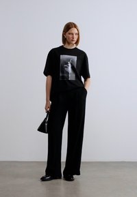 Μαύρο υπερμεγέθες μπλουζάκι με μονόχρωμο γραφικό, συνδυασμένο με μαύρο παντελόνι φαρδιάς γραμμής και μικρή μαύρη τσάντα χειρός.