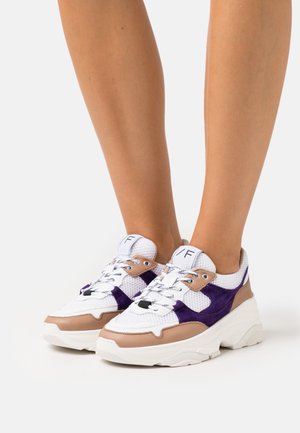 White sneakers with beige and purple accents worn on feet, featuring thick soles and white laces on a light background.