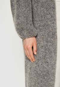 Main gauche reposant contre la cuisse, vêtue d'un pull en maille texturée grise à manches longues sur un fond clair uni.