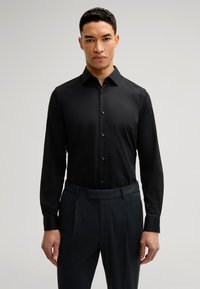 Zwarte langemouwen shirt met een knoopsluiting aan de voorkant, kraag en een zachte textuur, gecombineerd met donkere gestreepte broeken. Strakke pasvorm benadrukt de structuur.