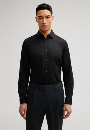 Black long-sleeve shirt with a button-up front, collar, and soft texture, paired with dark patterned trousers. Close-fit design highlights structure.