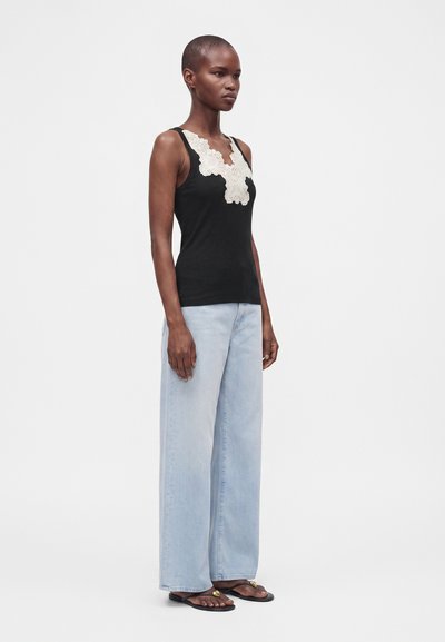Femme debout portant un débardeur noir avec des détails en dentelle blanche, un jean bleu clair à jambes larges et des sandales noires avec des accents dorés.