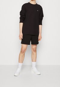Svart långärmad t-shirt med rund halsringning, kombinerad med svarta shorts. Vita sneakers och strumpor fulländar outfiten. Enkel, avslappnad design.