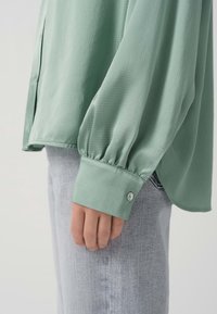 Camicia a maniche lunghe verde chiaro con superficie testurizzata, che crea una caduta morbida. Presenta un polsino con bottone e una vestibilità rilassata. Abbinata a jeans grigio chiaro.