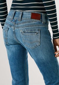 Jean en denim bleu délavé avec une coupe droite, comprenant un patch en cuir marron de la marque, des poches arrière et des coutures bien définies le long des coutures.