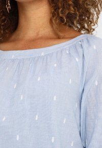 Lyseblå, off-shoulder bluse lavet af tekstureret stof med hvide ovale mønstre over det hele, med rynkede detaljer ved halsudskæringen.