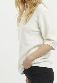 Maglione in maglia leggero di colore crema, con una vestibilità rilassata, maniche corte e dettagli a coste, indossato con pantaloni neri.