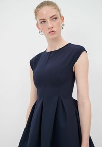 Laivastonsininen hihaton A-linjainen mekko, jossa on pyöreä pääntie, laskostettu hame ja siistit saumat. Sileä kangas, jossa on ryhdikäs siluetti.