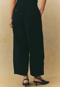 Pantaloni neri a gamba larga con un alto elastico in vita, dotati di due tasche posteriori con bottoni e una texture liscia e opaca.
