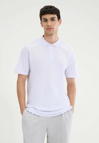 Biała koszulka polo z krótkim rękawem, regularny krój, wykonana z oddychającego materiału. Zauważalne wykończenie kołnierzyka i detale przy guzikach. Połączona z szarymi spodniami dresowymi.