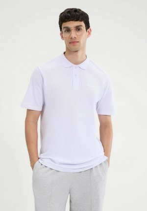 Weißes Poloshirt mit kurzen Ärmeln, normale Passform, aus atmungsaktivem Stoff. Auffälliger Kragen und Knopfdetails. Kombiniert mit grauen Sweatpants.