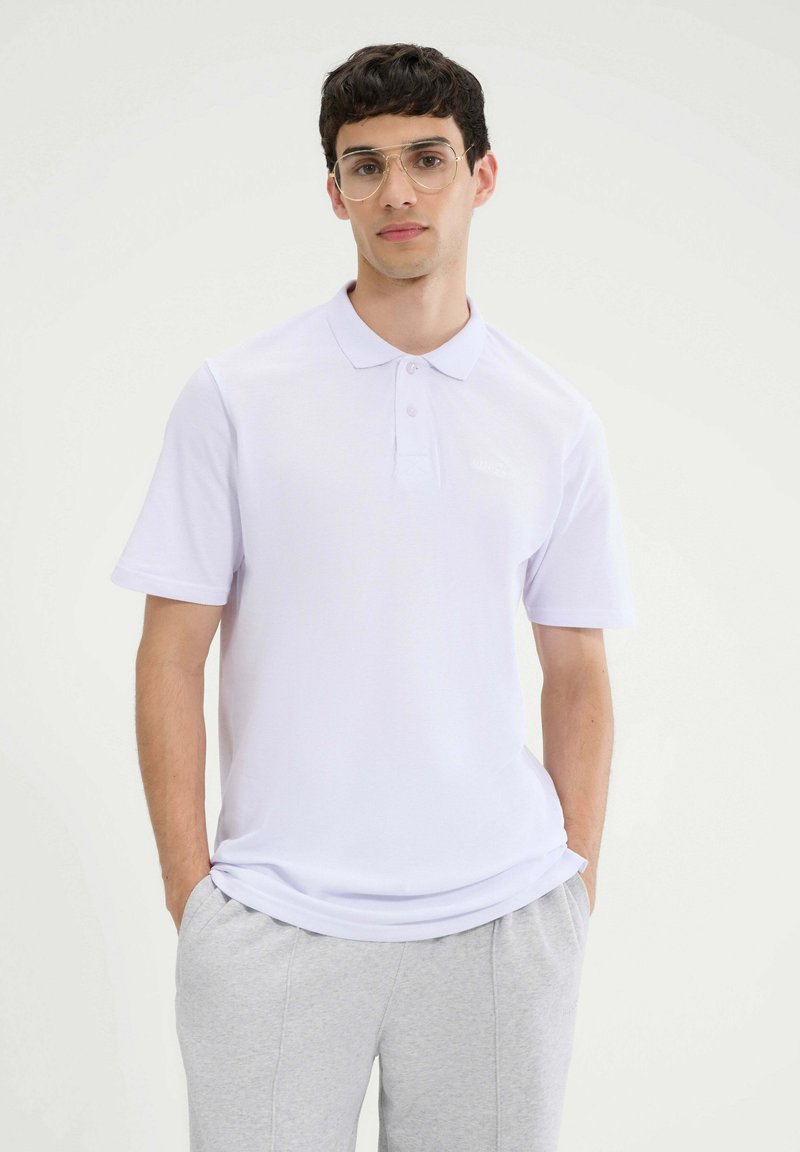 Biała koszulka polo z krótkim rękawem, regularny krój, wykonana z oddychającego materiału. Zauważalne wykończenie kołnierzyka i detale przy guzikach. Połączona z szarymi spodniami dresowymi.