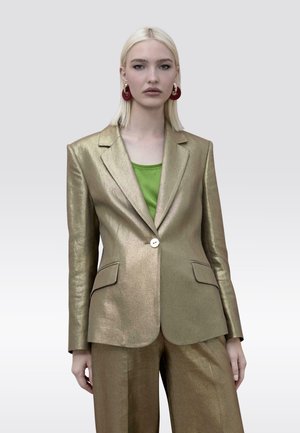 Mujer con cabello rubio lacio que lleva una chaqueta y pantalones metálicos dorados combinados con una blusa verde y grandes pendientes rojos circulares.