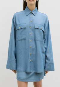 Helle blaue Jeanshemd mit spitzem Kragen, zwei Brusttaschen und Knopfleiste. Es hat lange Ärmel und einen lockeren Schnitt.