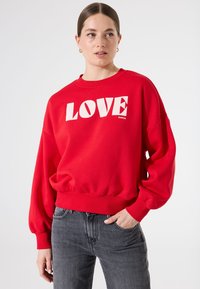 Sudadera roja con gráfico "LOVE" en blanco. Presenta un escote redondo y puños fruncidos, combinada con jeans grises.