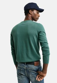 Grüner Strickpullover mit langen Ärmeln, rundem Halsausschnitt und gerippten Bündchen, kombiniert mit blauen Jeans. Lässige Passform, minimalistische Details.