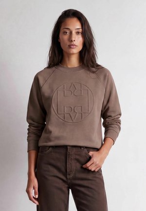 Femme aux longs cheveux foncés portant un sweat-shirt marron avec un logo circulaire en relief et un pantalon marron foncé, debout avec une main dans la poche.
