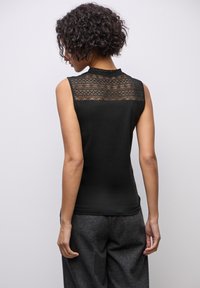 Top nero senza maniche con dettagli in pizzo sul retro, dotato di un colletto alto e una consistenza liscia. Abbinato a pantaloni grigi con una texture lavorata.