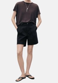 Camisa marrón de manga corta, pantalones cortos negros ajustados y sandalias negras con acentos de puntos. Atuendo minimalista y casual adecuado para el clima cálido.
