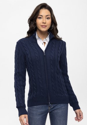 Cardigan lavorato a maglia blu navy con motivo a treccia, zip frontale completa, colletto alto, polsini e orlo a costine, abbinato a una camicia bianca e jeans.