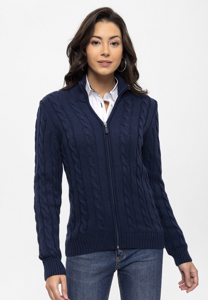 Marineblauwe gebreide cardigan met kabelpatroon, volledige rits aan de voorkant, hoge kraag, geribbelde manchetten en zoom, gecombineerd met een wit shirt en jeans.