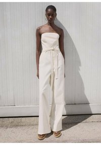 Combinaison blanche sans bretelles avec une taille ceinturée, une silhouette à jambes larges et une texture lisse. Le modèle se tient devant un fond en bois clair.