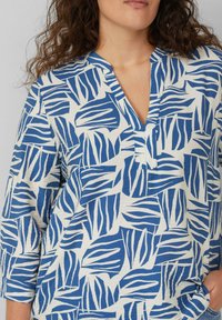 Blauw-witte top met een V-hals en driekwartmouwen. Heeft een bladerenpatroon op een gestructureerde stof. Voorkantzak zichtbaar.