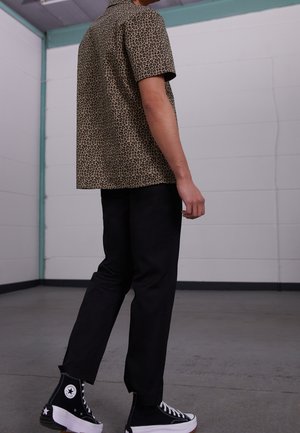 Kurzärmeliges Leopardenmuster-Hemd in Beigefarben mit schwarzem Muster, kombiniert mit schwarzen Hosen und schwarzen High-Top-Sneakern mit weißen Akzenten.