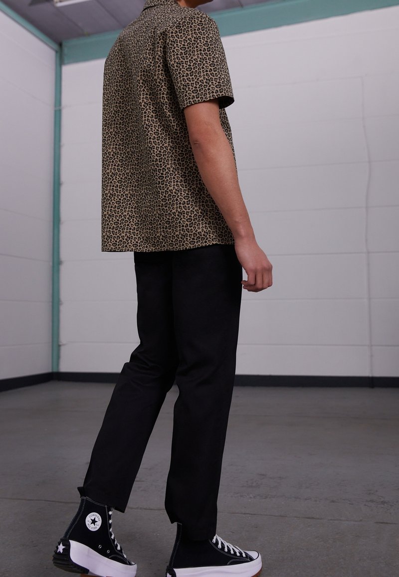 Koszula z krótkim rękawem w panterkę w odcieniach beżu i czerni, zestawiona z czarnymi spodniami i czarnymi wysokimi trampkami z białymi akcentami.