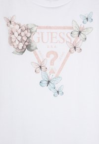 Valkoinen T-paita, jossa on kukkakuosi, joka esittää vaaleanpunaisia hortensioita ja sinisiä perhosia, painettu kolmion muotoon, jossa on "GUESS"-logo ruusukultaisena.