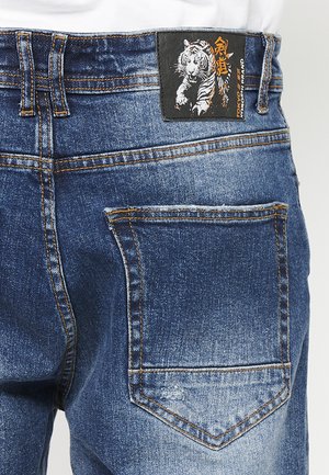 Blue denim jeans with contrasting golden stitching, featuring a back patch with a tiger graphic and a single back pocket.