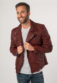 Giacca da motociclista rossa in pelle con tasche con zip, bottoni a pressione e colletto appuntito, indossata sopra una camicia grigia chiaro e abbinata a jeans blu.
