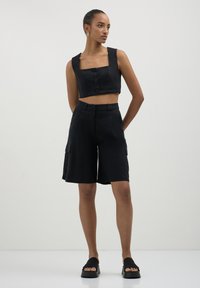 Donna in piedi con un top corto senza maniche nero con bottoni, pantaloni cargo alti fino al ginocchio e sandali neri con plateau.