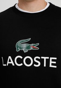 Černá mikina s šedým krokodýlem a nápisem "LACOSTE" silnými bílými písmeny. Límec má bílý lem.
