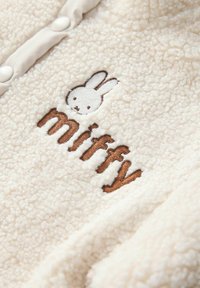 Kreemikatsega fliisteki, millel on broderi Miffy jänes ja sõna "miffy" pruunis värvuses, ees nupukinnitustega.