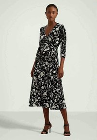 Mulher em pé vestindo um vestido midi envolvente preto com padrão floral branco e mangas três quartos, combinado com sandálias de salto pretas.