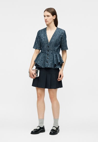 Femme portant un chemisier péplum texturé bleu marine avec des boutons noirs, une jupe plissée noire, des chaussettes grises, des mocassins noirs, et tenant une petite pochette argentée.