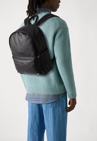 Mochila de cuero negro con una textura suave, bolsillo delantero con cremallera y correas ajustables, llevada sobre un suéter de punto azul claro y pantalones de mezclilla.