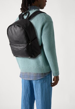 Mochila de cuero negro con una textura suave, bolsillo delantero con cremallera y correas ajustables, llevada sobre un suéter de punto azul claro y pantalones de mezclilla.