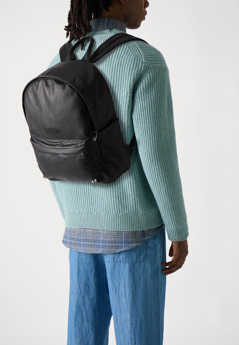 Mochila de cuero negro con una textura suave, bolsillo delantero con cremallera y correas ajustables, llevada sobre un suéter de punto azul claro y pantalones de mezclilla.