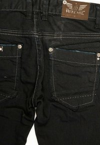 Jean en denim noir avec des coutures contrastantes, comprenant deux poches arrière, des rivets métalliques et une étiquette en cuir marron portant le nom "Ritchie".