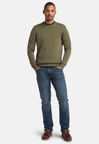 Maglione lavorato a maglia verde oliva con collo rotondo, abbinato a jeans denim blu e scarpe stringate marroni. Design casual e aderente.