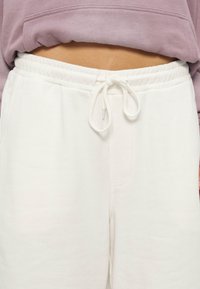 Μεσαίο τμήμα που δείχνει λευκό φόρμας με κορδόνι και ελαστική μέση, συνδυασμένο με μοβ κοντομάνικο φούτερ.