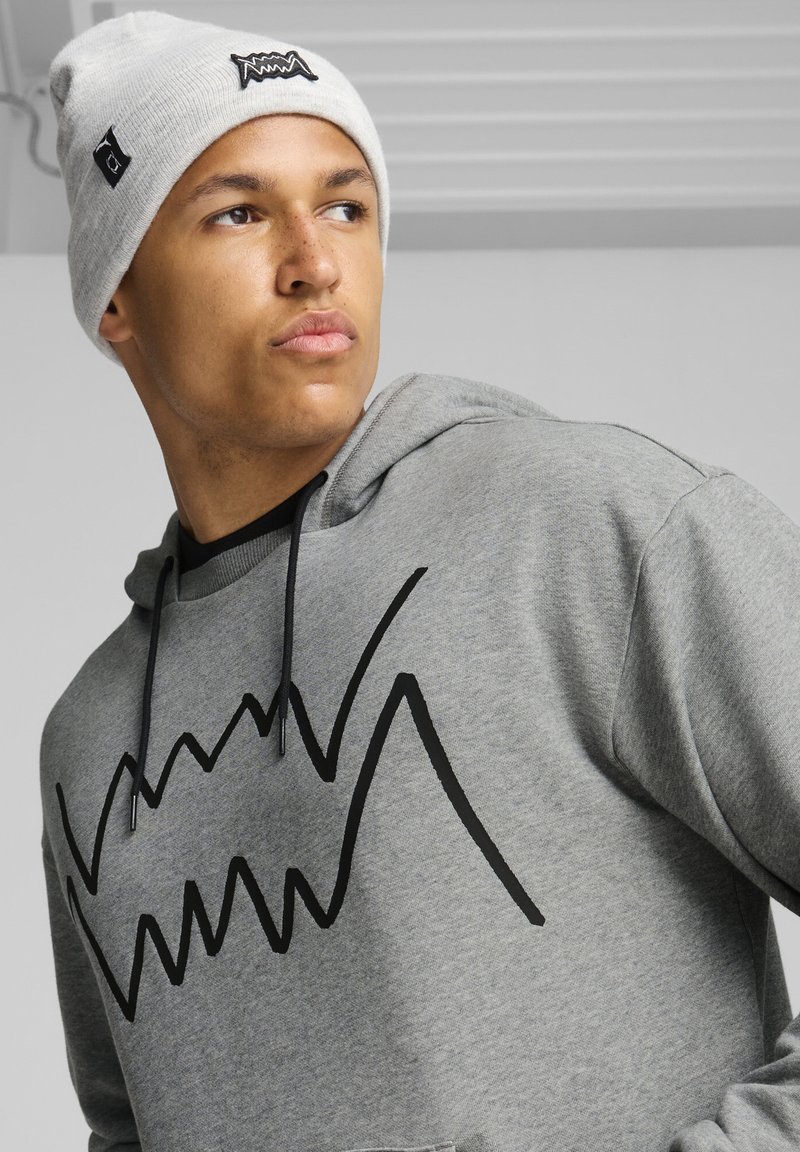 Haut à capuche gris avec un graphique en zigzag noir, doté d'une poche kangourou et d'un cordon de serrage. Porté avec un bonnet en maille gris clair affichant un logo.