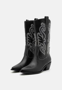 Bottes de cow-boy en cuir noir avec des pointes, broderies métalliques argentées et un talon empilé. Présentent des languettes pour un enfilage facile.