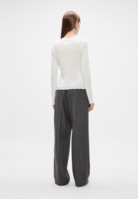 Bluză albă cu mânecă lungă și margine ondulată, asortată cu pantaloni largi gri cu dungi, prezentată din spate.