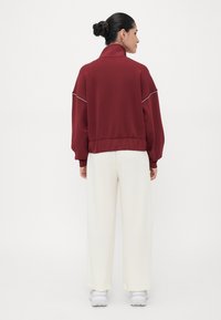 Burgunderfarget sweatshirt med høy krage og hvite detaljer, parret med løstsittende hvite bukser og hvite sneakers, sett fra bak.